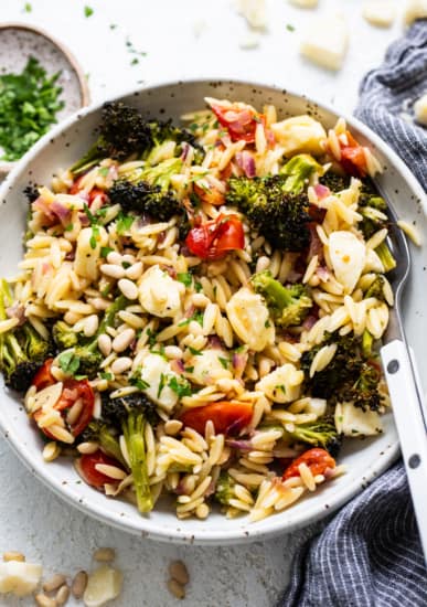 A bowl of orzo salad with roasted broccoli, tomatoes, and pine nuts, sprinkled with cheese and herbs, on a textured white surface.