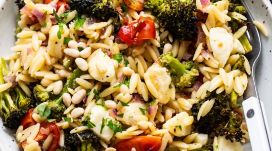 A bowl of orzo pasta salad with roasted broccoli, cherry tomatoes, and pine nuts, garnished with herbs.