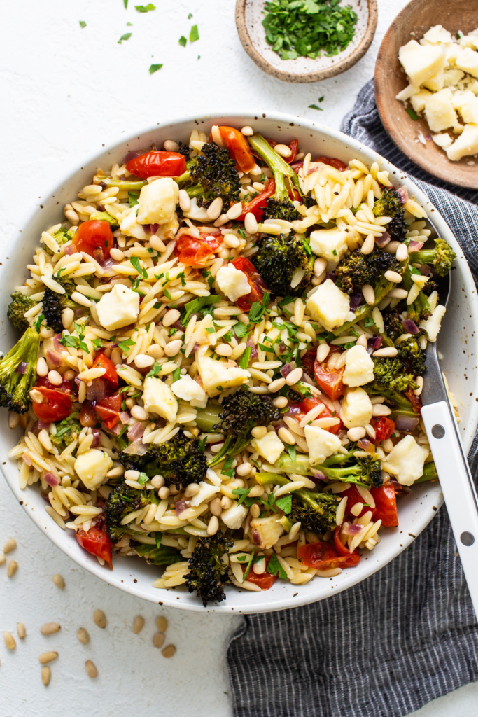 A vibrant orzo salad with broccoli, tomatoes, and pine nuts in a white bowl, garnished with herbs and cheese, on a textured white surface.