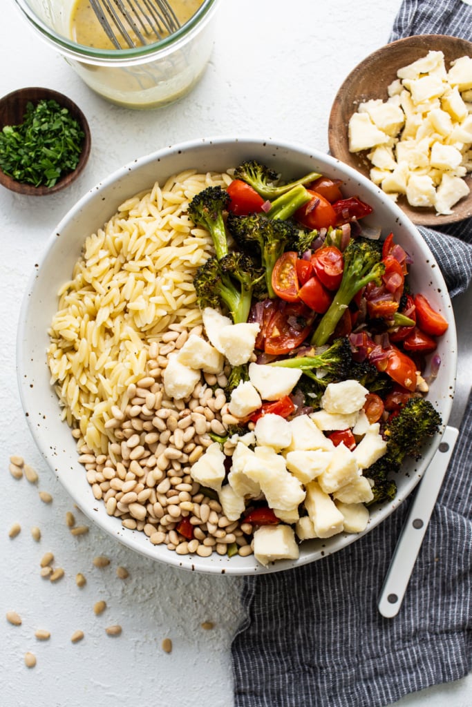 A bowl of salad with orzo pasta, roasted broccoli, cherry tomatoes, mozzarella cheese, and pine nuts on a white surface.