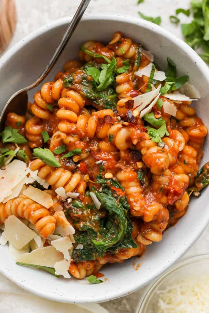 A bowl of fusilli pasta mixed with tomato sauce, spinach, and topped with shaved parmesan cheese.