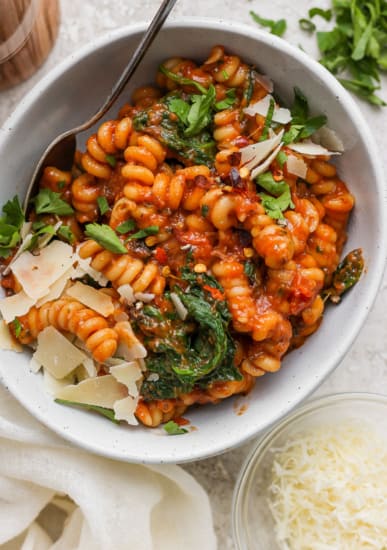 Bowl of tomato-based pasta with spinach and shaved parmesan cheese.