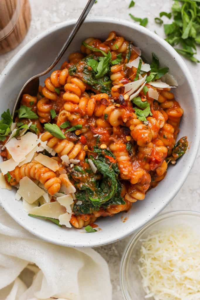 Bowl of tomato-based pasta with spinach and shaved parmesan cheese.