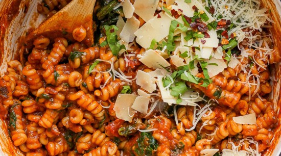 A pot of tomato-based pasta with spinach, garnished with grated cheese and herbs.