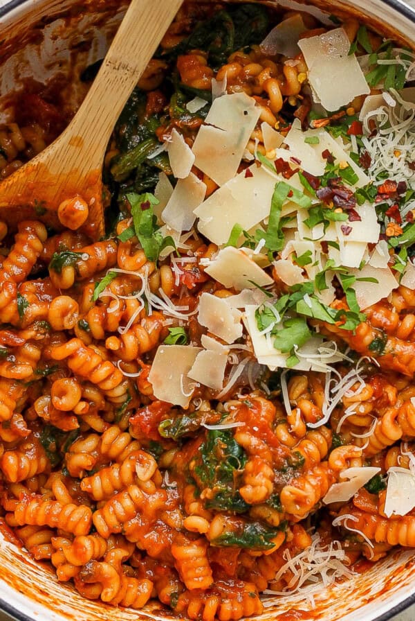 A pot of tomato-based pasta with spinach, garnished with grated cheese and herbs.