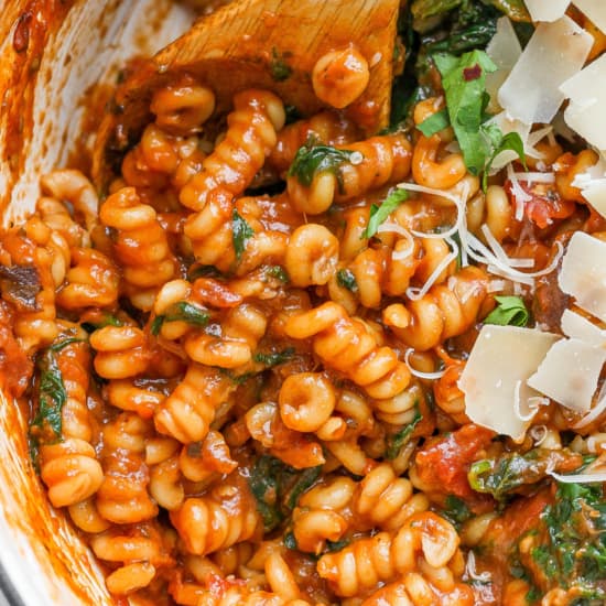A close-up image of a pasta dish with tomato sauce, garnished with spinach and sprinkled with shredded cheese.