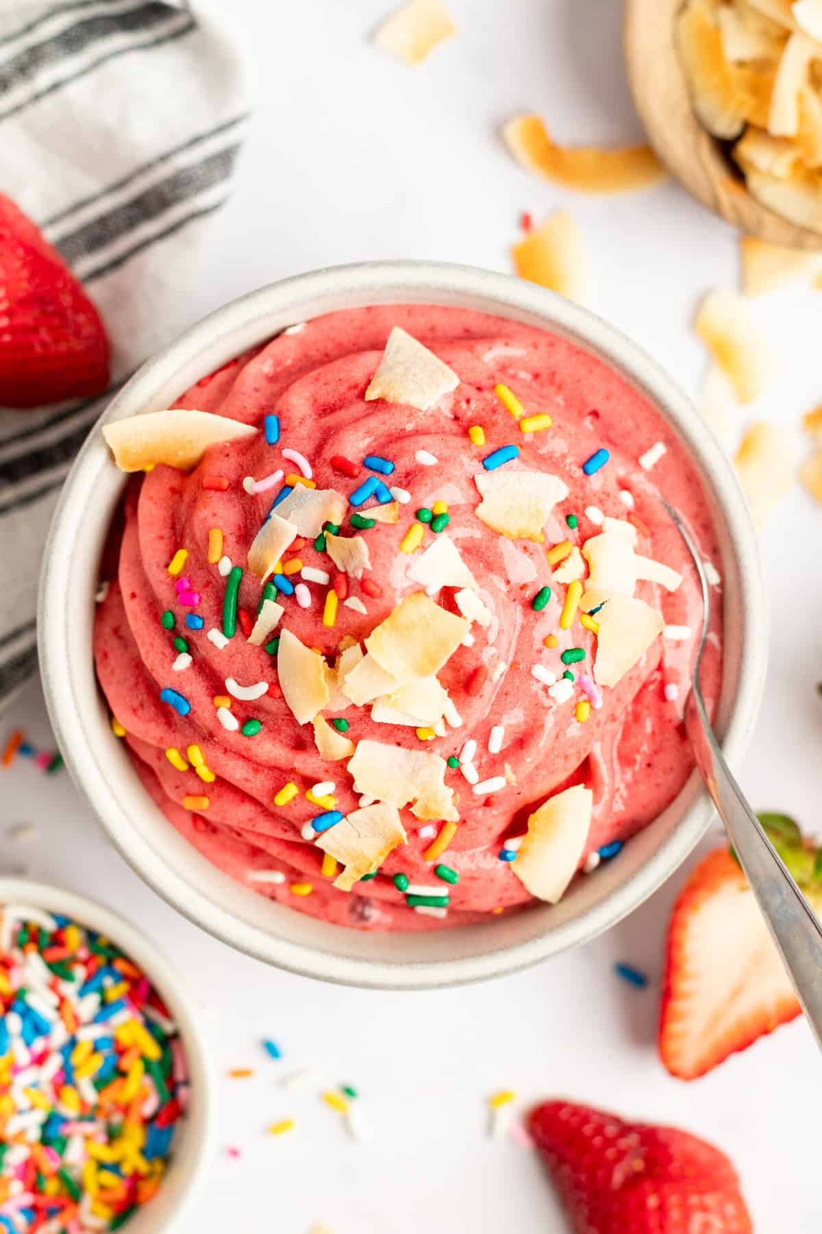 A vibrant pink smoothie bowl topped with coconut flakes and rainbow sprinkles, accompanied by fresh strawberries and a spoon.