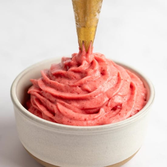 A piping bag dispensing creamy strawberry frosting into a small beige bowl against a white background.