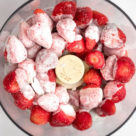A food processor bowl filled with whole strawberries and scoops of vanilla ice cream, viewed from above.