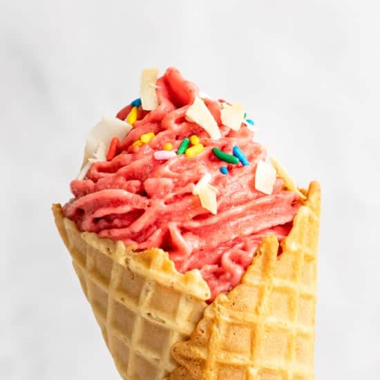 A close-up of a strawberry soft serve ice cream cone topped with sprinkles and white chocolate chunks.