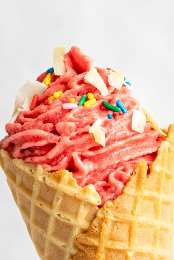 A close-up of a strawberry soft serve ice cream cone topped with sprinkles and white chocolate chunks.