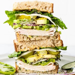 A tall sandwich with multiple layers of turkey, cheese, lettuce, cucumber, avocado, and sprouts on whole grain bread, served on a ceramic plate.