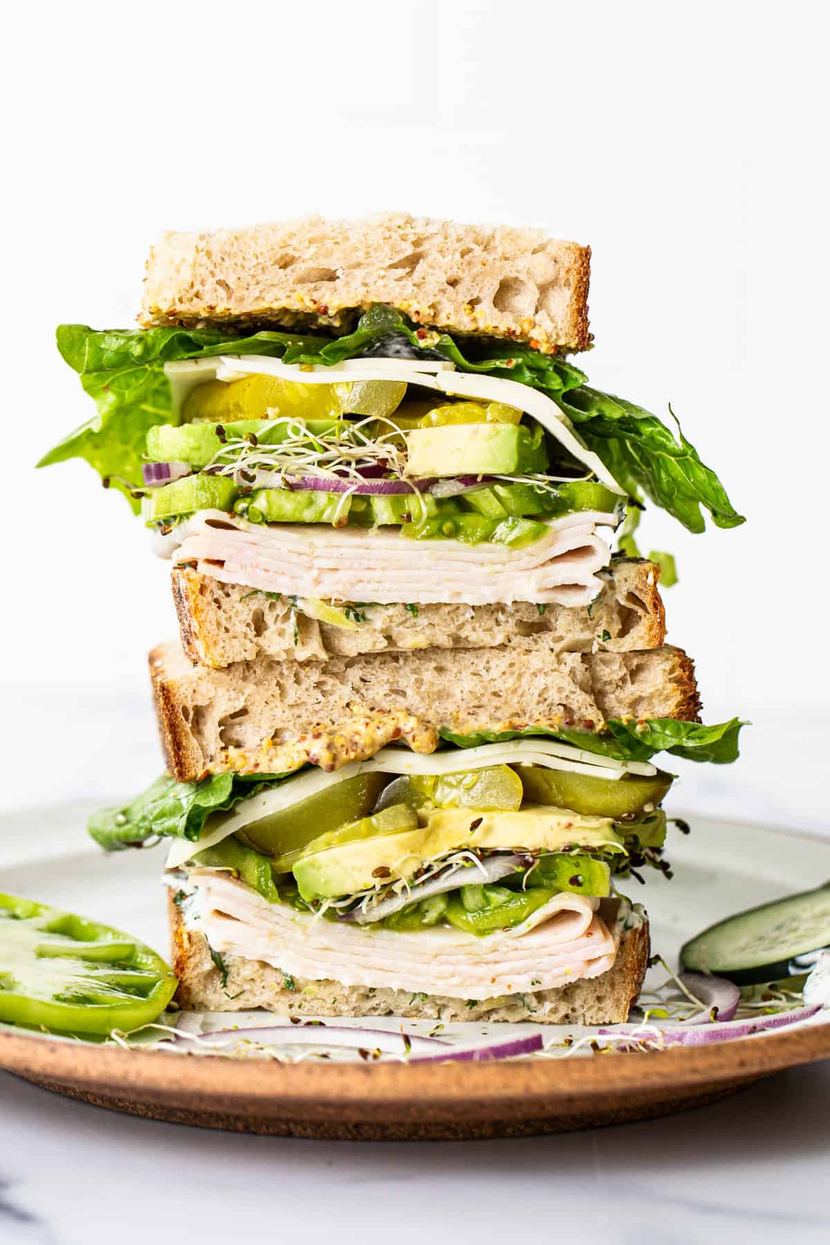 A tall sandwich with multiple layers of turkey, cheese, lettuce, cucumber, avocado, and sprouts on whole grain bread, served on a ceramic plate.