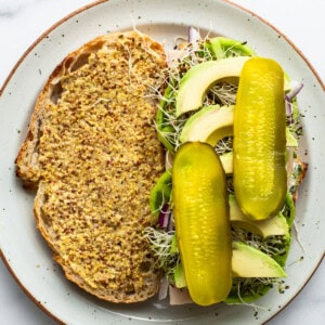 A plate of open-faced sandwich with mustard-covered bread, sprouts, sliced avocado, and two whole pickles on top.