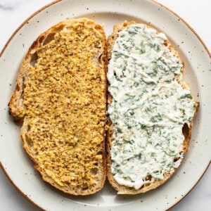 Two slices of bread on a plate, one topped with mustard and herbs, the other with a creamy white spread and chopped dill.