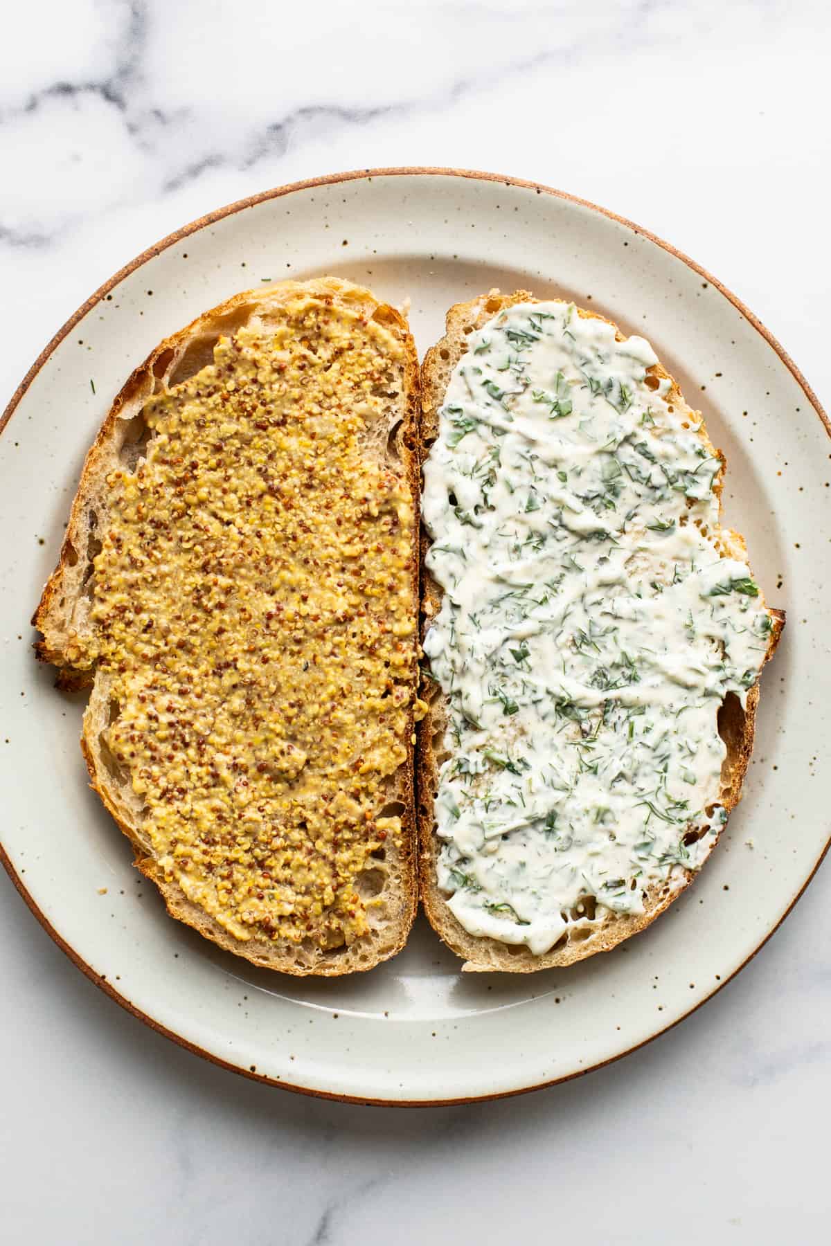 Two slices of bread on a plate, one topped with mustard and herbs, the other with a creamy white spread and chopped dill.