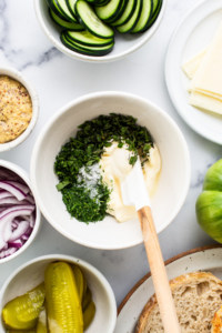 Overhead view of a kitchen counter with ingredients for a sandwich, including sliced cucumbers, cheese, pickles, red onions, and a bowl of mayonnaise mixed with herbs.