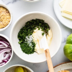 Overhead view of a kitchen counter with ingredients for a sandwich, including sliced cucumbers, cheese, pickles, red onions, and a bowl of mayonnaise mixed with herbs.