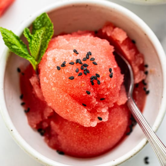A bowl of watermelon sorbet garnished with mint and black sesame seeds, served with a s،.