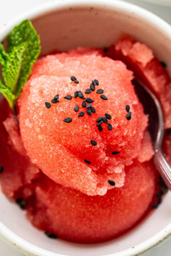 A bowl of watermelon sorbet garnished with mint and black sesame seeds, served with a spoon.