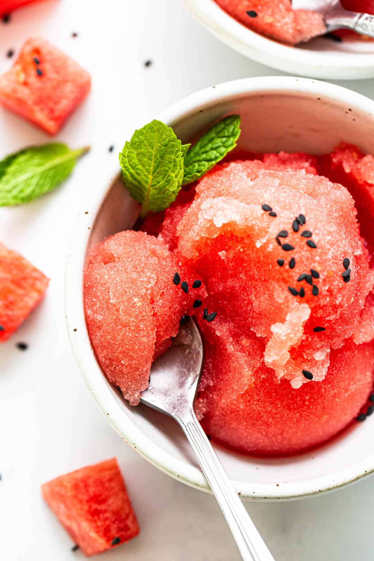 Bowl of watermelon sorbet garnished with mint leaves and black sesame seeds, with a spoon and scattered watermelon pieces around on a white background.