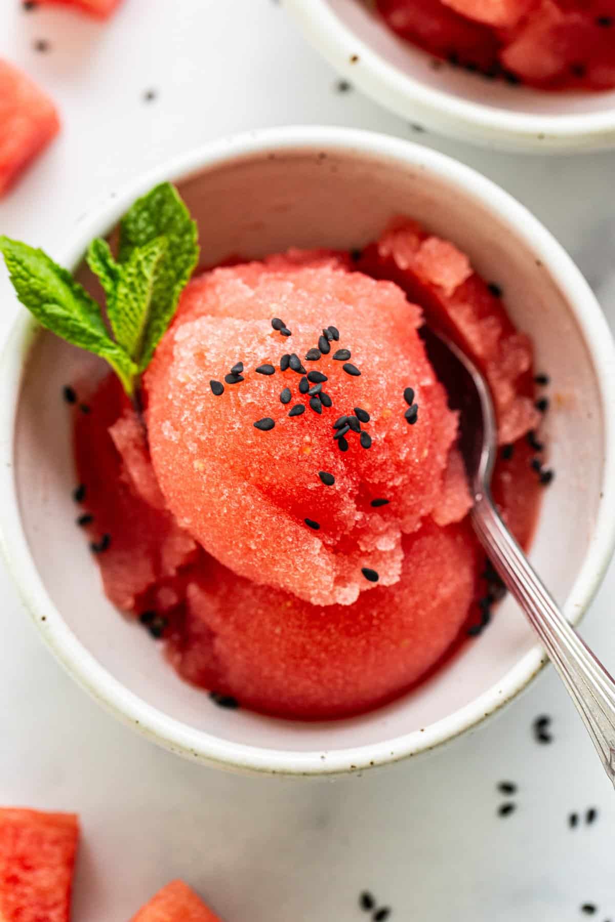 A bowl of watermelon sorbet garnished with mint leaves and black sesame seeds, accompanied by a spoon, on a light surface.