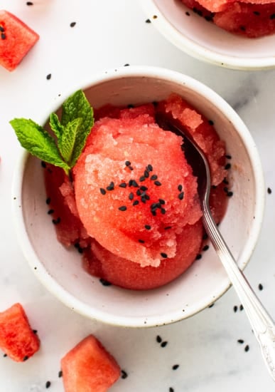 A bowl of watermelon sorbet garnished with mint and black sesame seeds, with a spoon, on a white surface scattered with diced watermelon pieces.