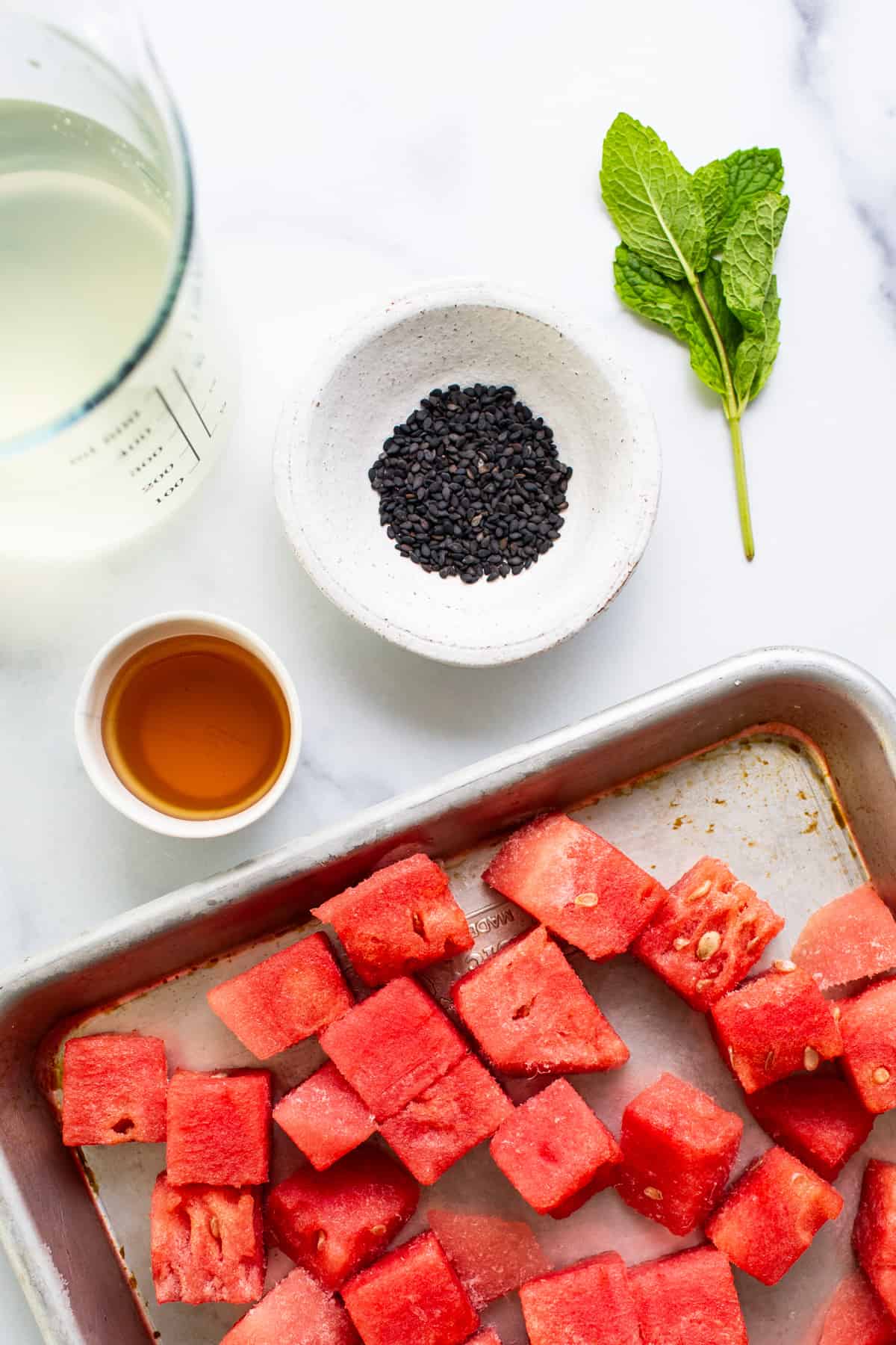 A tray of diced watermelon, a small bowl of black sesame seeds, a sprig of mint, a container of honey, and a glass of a pale beverage on a white surface.