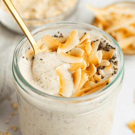 A glass jar filled with creamy coconut latte overnight oats, topped with toasted coconut flakes and chia seeds, with a gold spoon inserted.