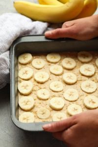 Person placing sliced bananas onto banana baked oatmeal batter in a baking tray, with whole bananas and a cloth in the background.