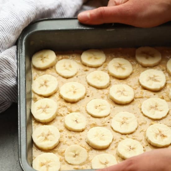 Person placing sliced bananas onto banana baked oatmeal batter in a baking tray, with w،le bananas and a cloth in the background.