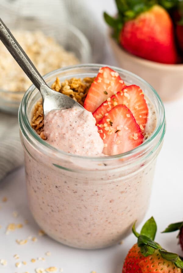 A jar filled with strawberry yogurt topped with granola and fresh sliced strawberries, with a spoon dipped in. A bowl of oats and whole strawberries are in the background.