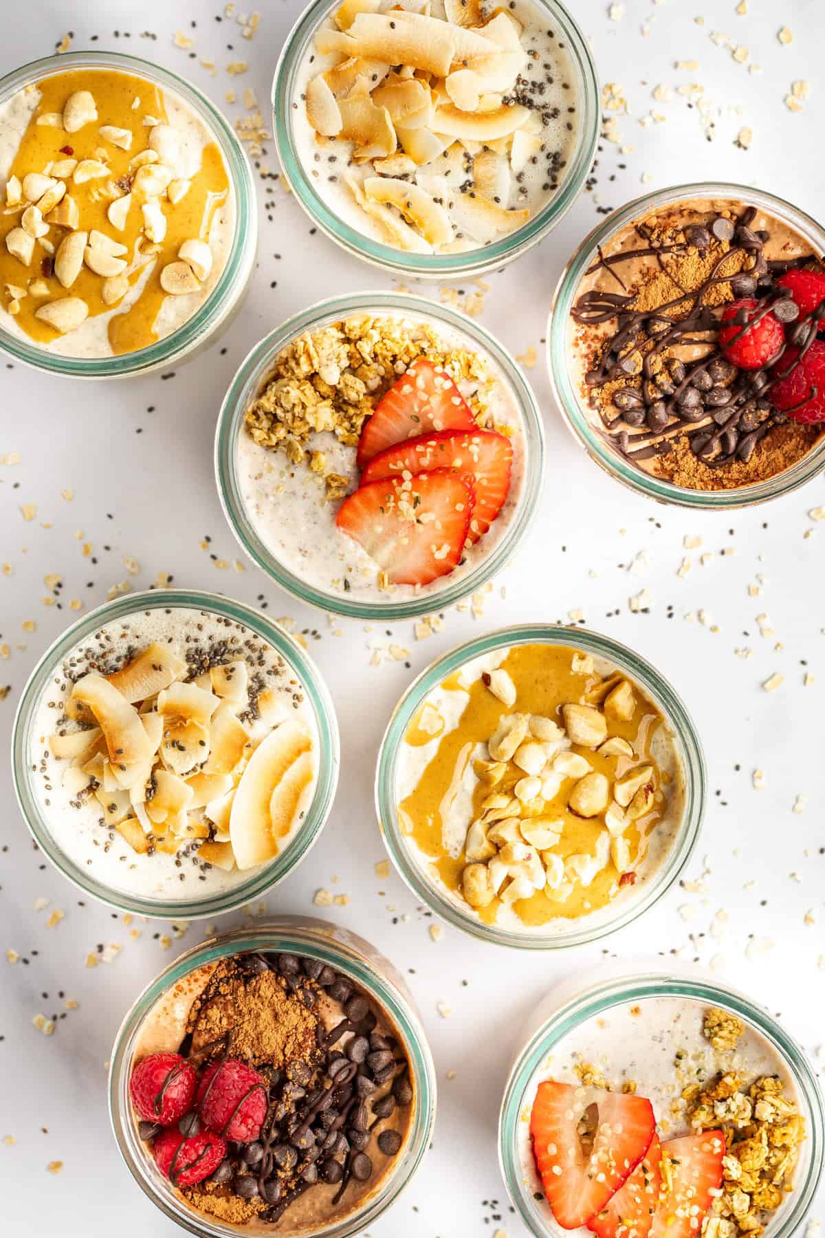Overhead view of seven jars filled with different varieties of overnight oats topped with ingredients like strawberries, chocolate chips, chia seeds, coconut flakes, and granola, arranged in a pattern.