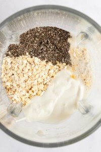 Ingredients including chia seeds, oats, and a creamy liquid mixture in a blender container, viewed from above.