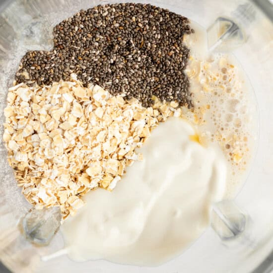Ingredients including chia seeds, oats, and a creamy liquid mixture in a blender container, viewed from above.