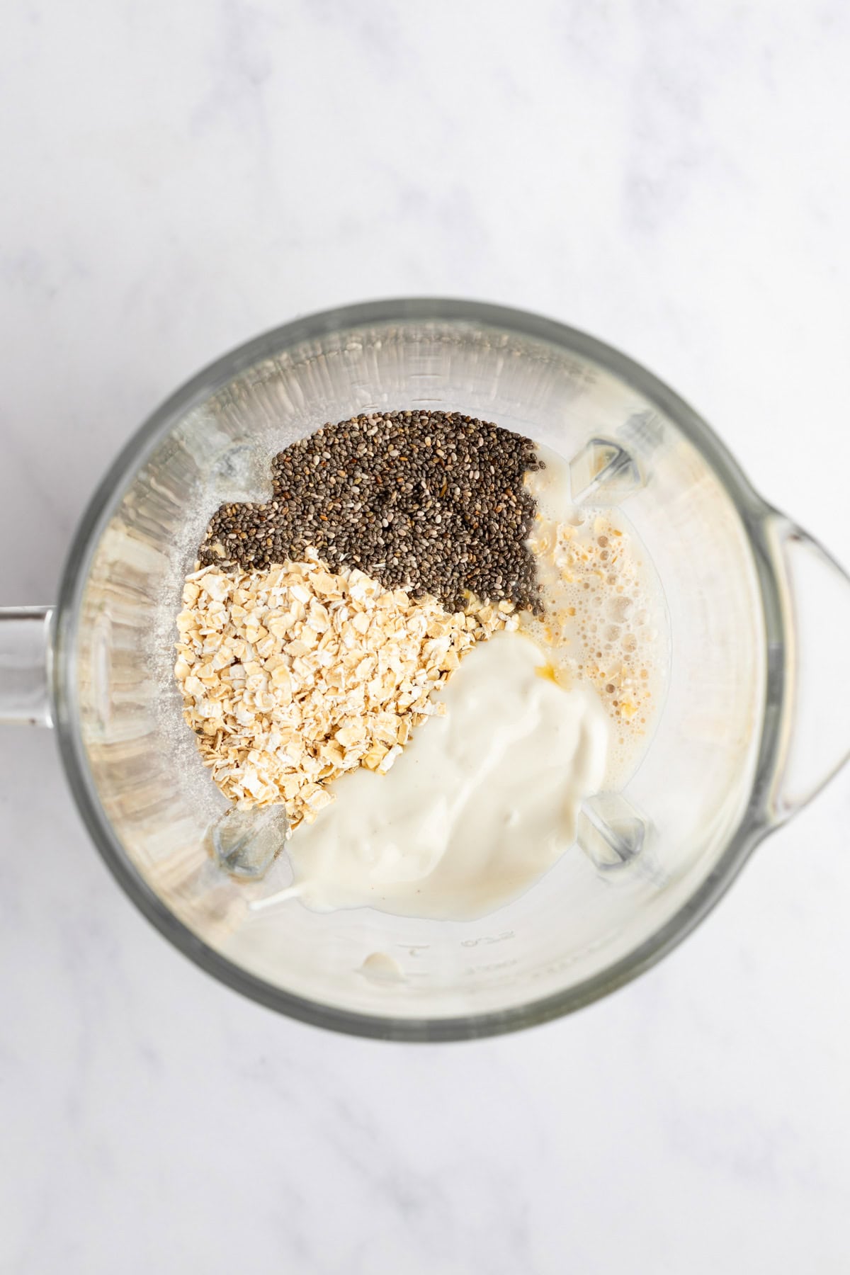 A blender containing chia seeds, oats, a white creamy substance, and liquid viewed from above on a light-colored surface.