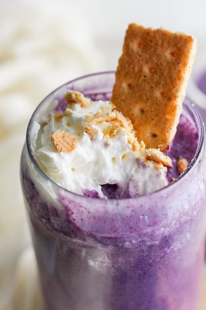 A close-up of a purple milkshake topped with whipped cream, crumbled cookies, and a cookie sticking out, served in a clear glass.