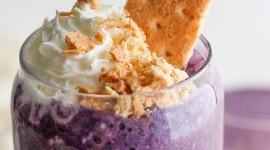 A creamy purple smoothie topped with whipped cream and crumbled biscuits, accompanied by a whole biscuit.