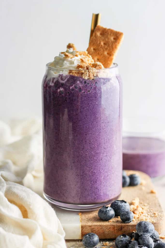 A vibrant purple smoothie topped with whipped cream and a sprinkle of crushed biscuits, served in a glass jar with fresh blueberries and a biscuit on the side.