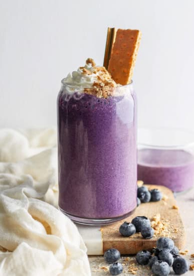 A vibrant purple smoothie topped with whipped cream and a cookie, garnished with blueberries on a light, textured background.