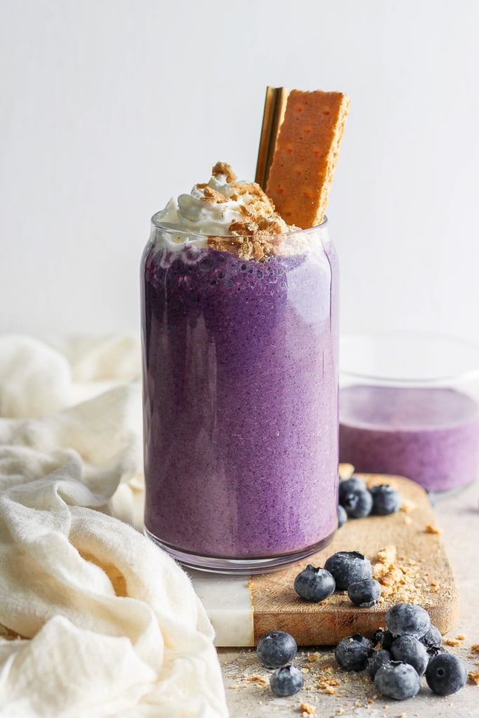 A vibrant purple smoothie topped with whipped cream and a cookie, garnished with blueberries on a light, textured background.
