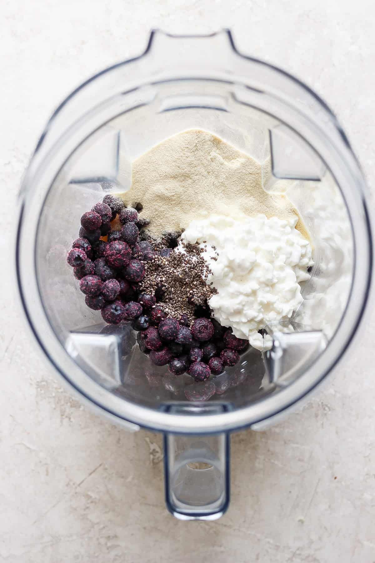 Top view of a blender containing blueberries, yogurt, chia seeds, and protein powder on a light surface.