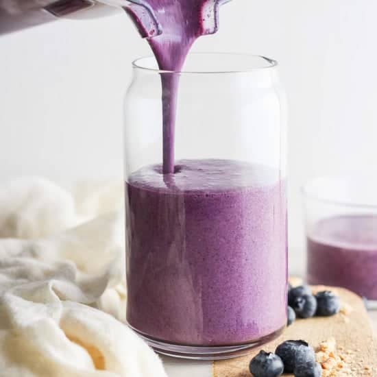 Pouring a fresh blueberry smoothie into a glass jar, surrounded by scattered blueberries on a wooden board.