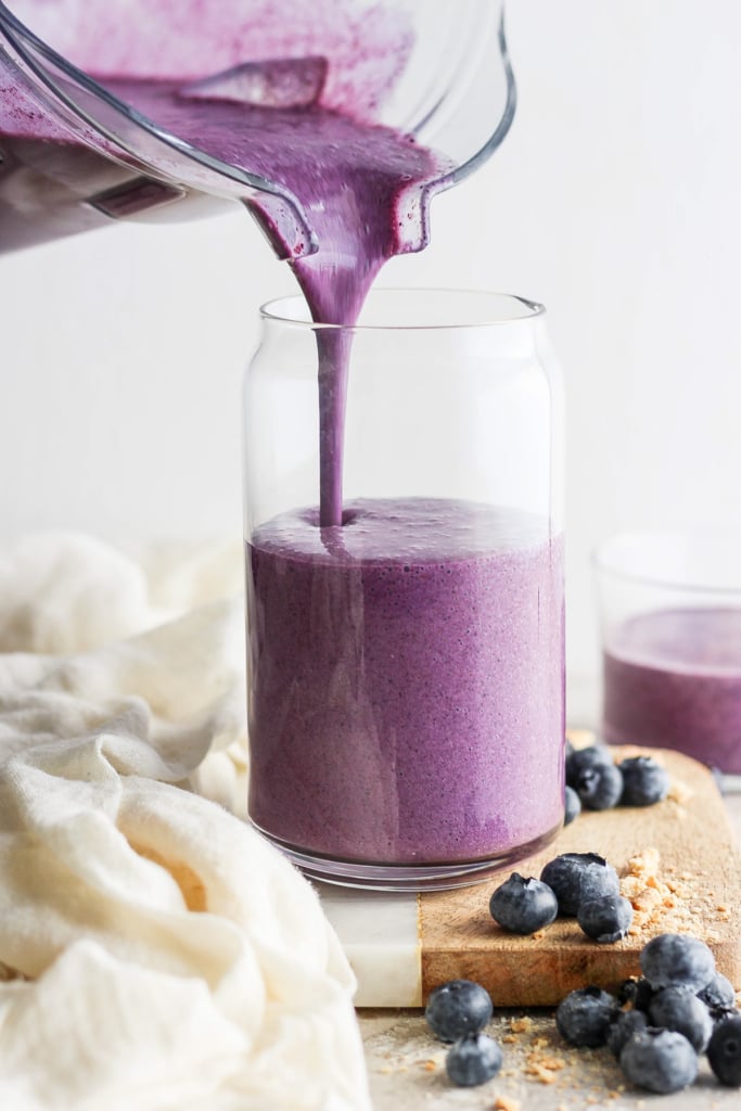 Pouring a fresh blueberry smoothie into a gl، jar, surrounded by ،tered blueberries on a wooden board.