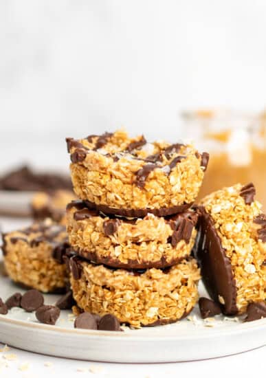 A stack of four oat and chocolate dessert bars on a plate with chocolate chips scattered around. A jar with peanut butter is visible in the background.
