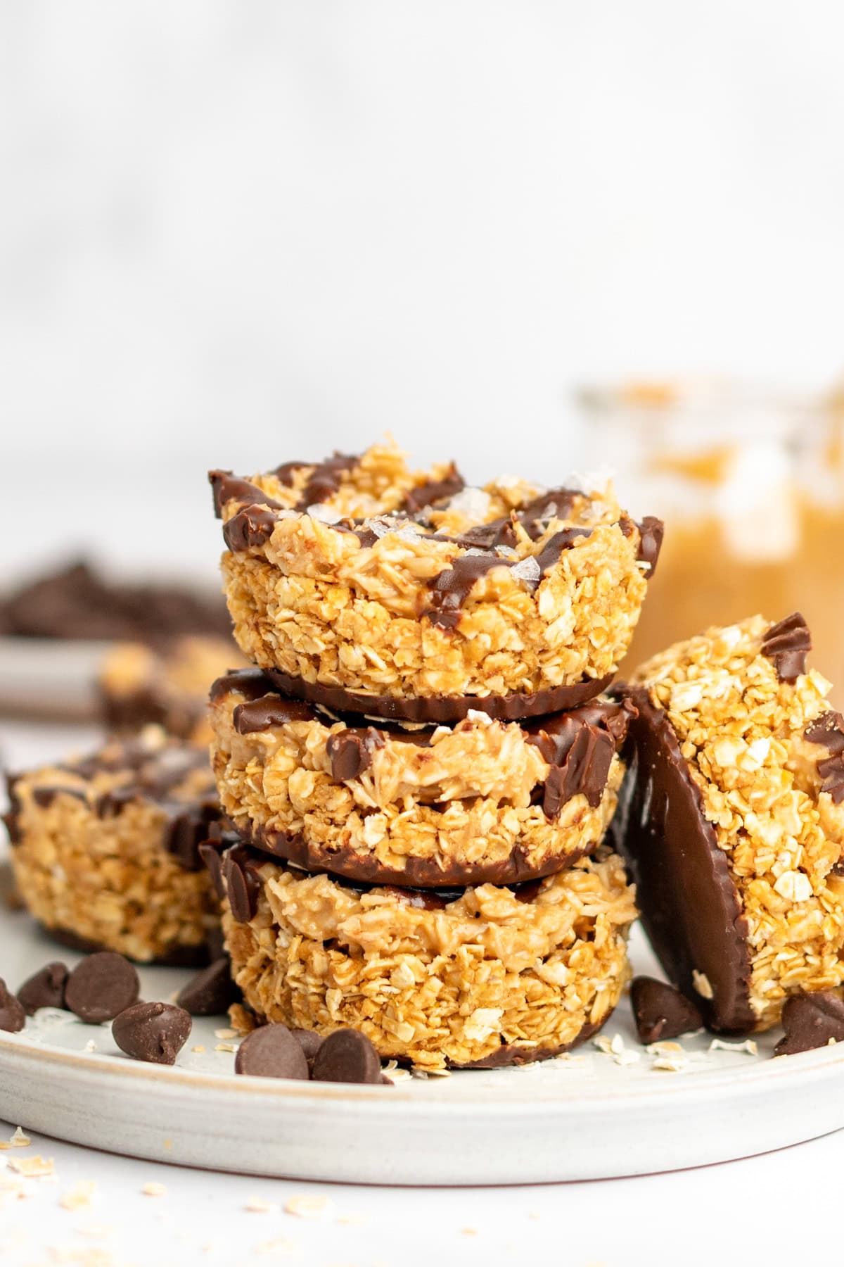A stack of four oat and chocolate dessert bars on a plate with chocolate chips scattered around. A jar with peanut butter is visible in the background.