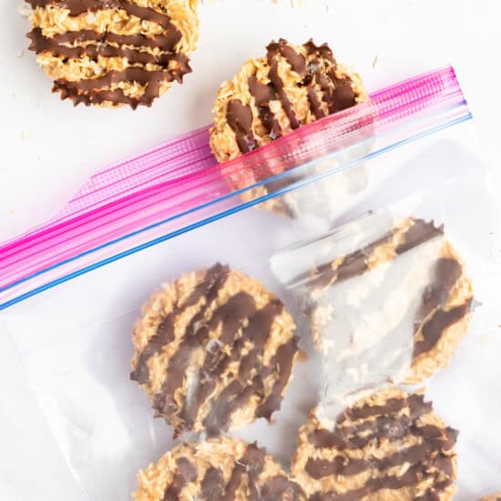 A clear plastic bag with a pink zip seal contains round cookies drizzled with chocolate. Two cookies are placed outside the bag.