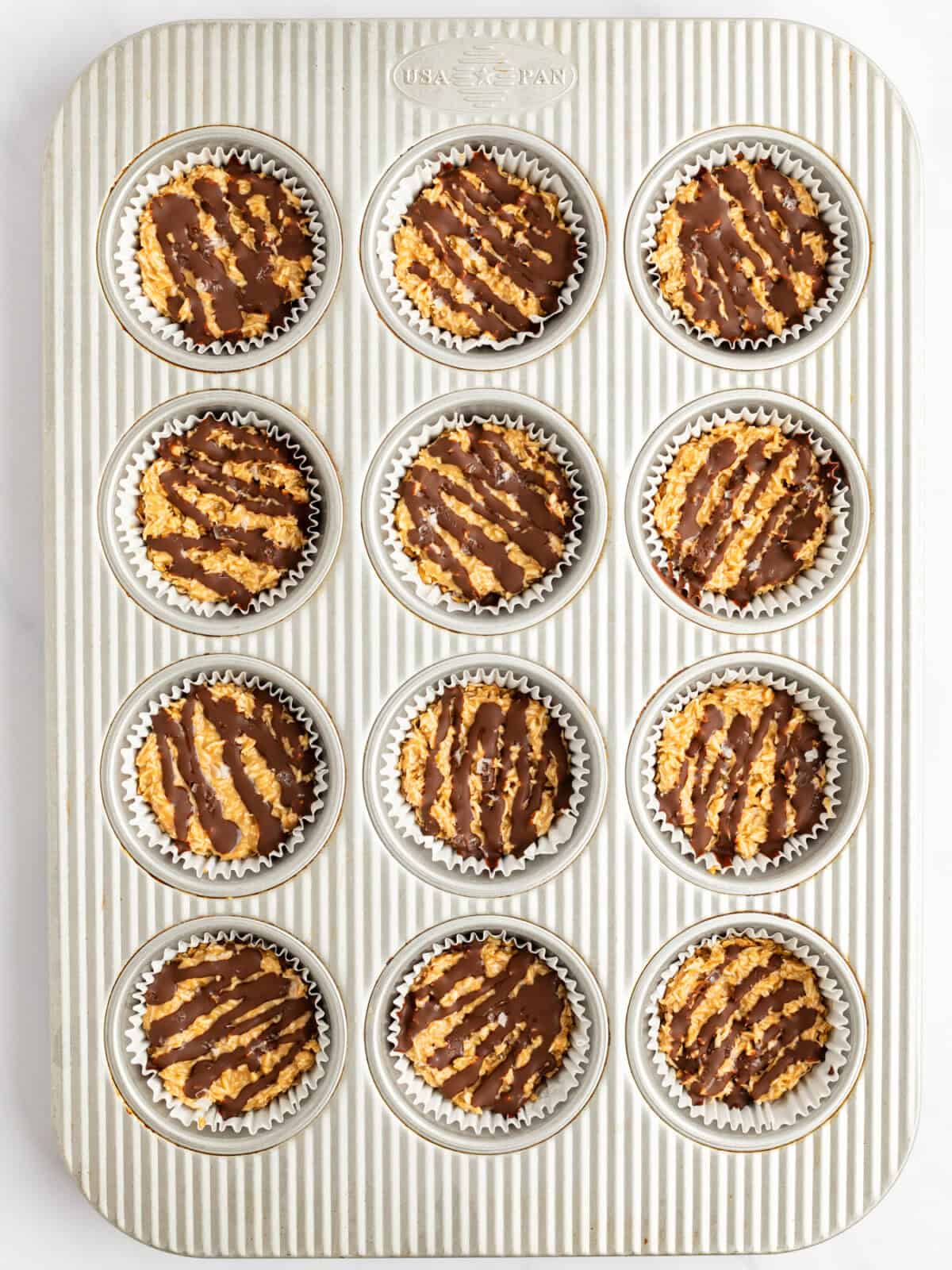 A muffin tin filled with 12 chocolate-drizzled oatmeal cookies in white paper liners.