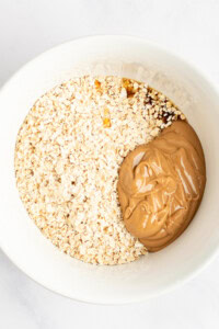 A white bowl containing a mixture of oats, peanut butter, and a brown liquid ingredient, possibly honey or syrup, inside.