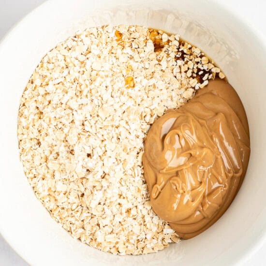 A white bowl containing a mixture of oats, peanut butter, and a brown liquid ingredient, possibly honey or syrup, inside.